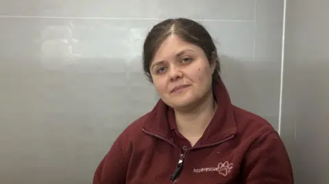 Hope Rescue Sara Rosser pictured sitting against a wall wearing burgundy Hope Rescue branded fleece. She has long dark hair tied back in a pony tail.