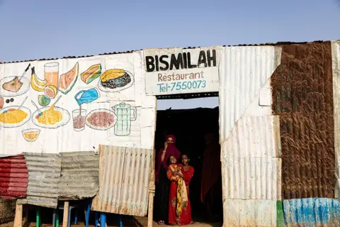 Aliona Sinenko/ICRC A woman and two children are seen standing in the doorway of a cafe in the Garowe refugee camp.