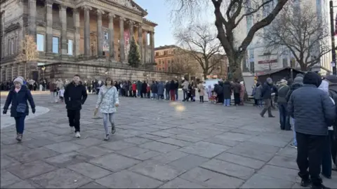 A queue with dozens of people outside a building with columns on the outside. They are standing on large stone flags, and the queue passes by trees without leaves. they are dressed for winter weather.