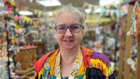Sonya Ellis is wearing multi-coloured glasses, green patterned dungaree, a multi-coloured top and a yellow lanyard with bees on it and is standing in her shop with a blurred background of craft items.