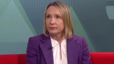 A woman with long blonde hair, a white shirt and purple jacket, speaks while looking to her right. She is sitting on a red sofa in a TV studio.