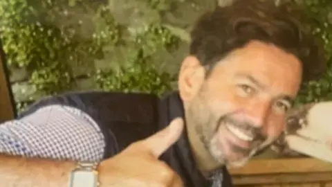 Merseyside police Martin O'Donovan smiles as he gives the camera a thumbs up. He has dark hair and a short beard. He wears a checked shirt under a black bodywarmer