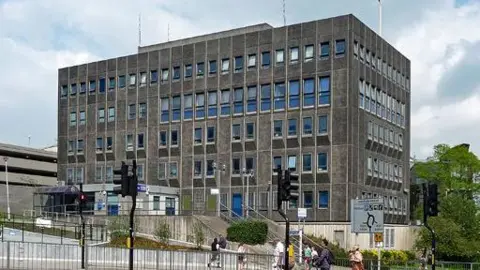 Stephen Richards/Geograph Charles Cross Police Station in Plymouth. It is a large multi-storey building near a roundabout. It is rectangular - cuboid shaped. Several people are crossing the road near the station.