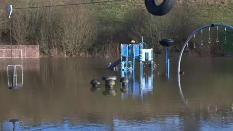 Falstone Janet/ Weather Watchers A playground submerged in water so much so that you cannot see any grass. You can see a blue slide set and monkey bars with the reflection from the water.