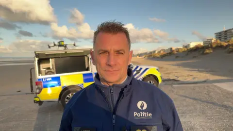 Jack Valpy/BBC Christian De Ritter, the police chief, standing on a beach in front of a police vehicle and a drone