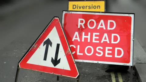 Getty Images Three road signs, one triangular with black arrows pointing up and down, one red with the words road closed on it and a smaller yellow sign which reads diversion