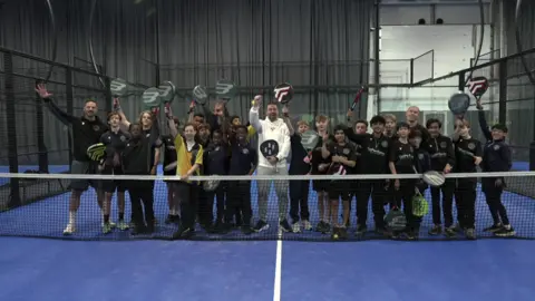 A picture of Jason Manford, dressed in a white top, stood behind a padel net with children either side of him, with them all cheering.