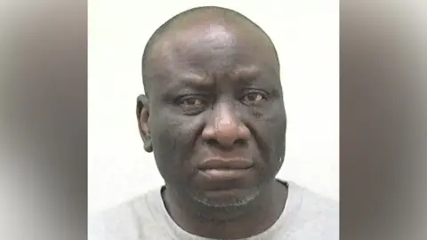 A police mug shot of Matthew Bello. He has a bald head and stubble on his chin. He is wearing a grey T-shirt.