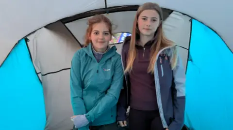 Two girls standing side-by-side in a tent. They are wearing coats and outdoor gear.