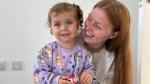 BBC Jacqueline Lowry and her daughter Keely Rose both smiling for the camera. Keely Rose is wearing a jumper with cartoons on it. Jacqueline is looking at her, she has red hair and is wearing a white t-shirt.