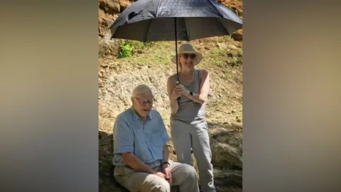 Chris Watson David Attenborough on the set of a documentary, sitting on a duty bank, sheltering in the shade of an umbrella held by a woman. Sir David is wearing a light blue shirt and beige trousers. The woman is wearing a sunhat, sun glasses, a grey vest and beige trousers.