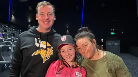 Two adults and a small child are seen in a movie theatre.
Faye is wearing a bright pink Derry City top and black club hat.
Her two parents stand either side of her and are both smiling.