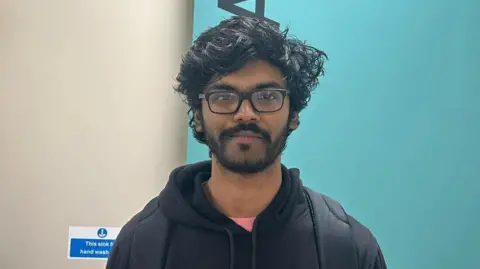 BBC Muphuselban stands in front of a blue background. He has black hair and black glasses. He has a black moustache and beard. He is wearing an orange top with a black hoodie.