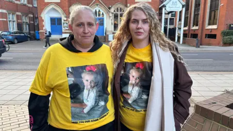 Two women standing together. They both have blonde hair and are wearing bright yellow T-shirts with a picture of a toddler in a pink hair bow. The text on the T-shirts reads "Miss you our angel Viviana-Ray".
