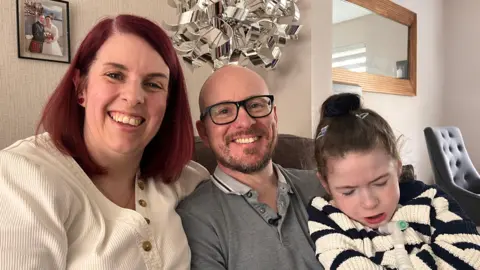 Family photo of three white people. Red haired mum Lynsey, age 44, with cream v-neck jumper sitting beside her husband Mark, age 46, who is bald with a beard and thick black glasses wearing a gray polo neck top. He is holding his daughter Thea who is wearing a black and cream striped jumper. Thea has a breathing and feeding tube coming out from her neck. They are sitting on a couch in a living room with a sliver lamp and wedding photo in the background.