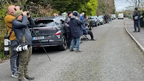 Garry Wilkinson People and cars lined along the road, people have cameras and are looking through binoculars 