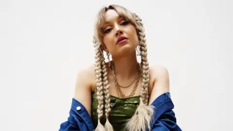 Rachel Jane Promo shot of Rachel Jane who has long blonde plaits and a shirt hanging off her shoulders