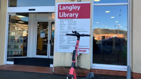 The frontage of Langley Library, with double doors and large windows, an opening times sign, and a scooter parked outside.