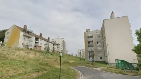 Two blocks of flats. The flats are cream covered with brown dirt marks on the exterior. They are at the end of the road at the top of a small grassy hill.