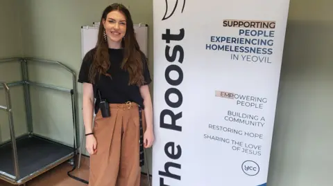 Bridget Doney stood against a white sign which reads 'The Roost. Supporting people experiencing homelessness in Yeovil. Empowering people, building a community, restoring hope and sharing the love of Jesus'. Bridget is smiling into the camera.