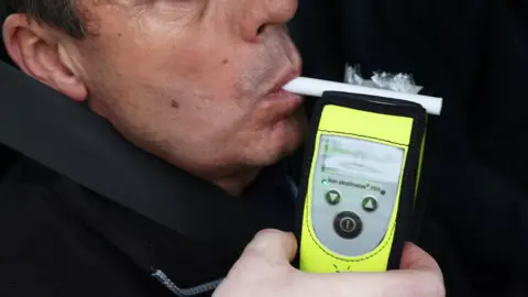 A man being breathalysed. It is a close-up shot, cropping out the top half of his face. He is blowing into a small white tube which is attached to the hand-held yellow breathalyser unit. The unit is held by someone who appears to be stood behind the camera.