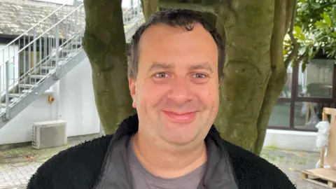 A smiling man in a grey T-shirt and fleece stands in front of a tree.