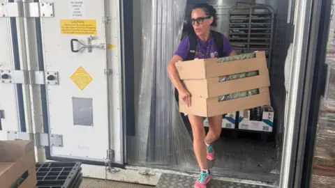 Towcester Community Larder A woman, wearing shorts and a purple T-shirt, with a backpack on her back, walks out of a cold storage unit holding three boxes of food. She has dark hair tied back and is wearing glasses.