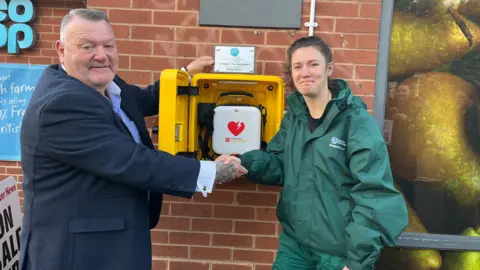 A man in a navy blue shirt with a tiger tattoo on his right hand is shaking a woman with brown hair in a ponytail and dressed in a green paramedic jacket. She is slightly smiling. Both are stood next to an opened  yellow box fixed on a red brick wall. there is a small white bag with a red heart printed on it inside.