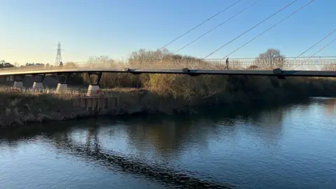 A long bridge with railings on either side crosses a river on a bright day. A person is crossing the bridge on foot.