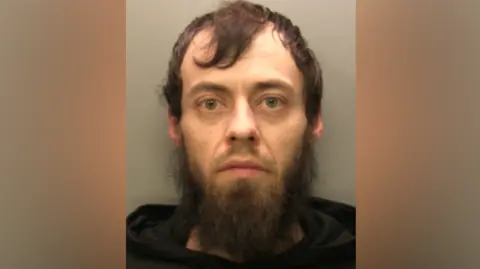 Jamie Hardy stands in front of a grey screen. He has a long black beard and long black hair and is wearing a black hoodie.