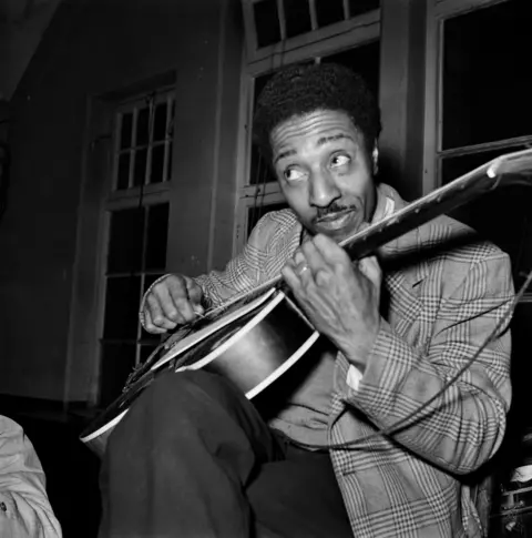 Bert Hardy/Getty Images A man wearing a chequered jacket and dark trousers plays guitar.