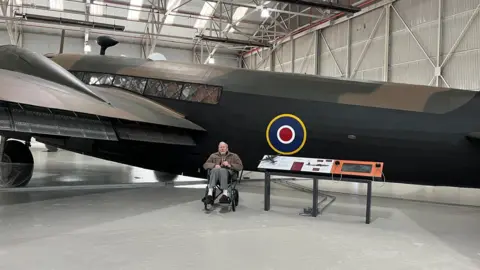 Supplied Donald Merryweather sitting in front of a Wellington bomber at the museum. He is in a wheelchair next to an information board about the aircraft, which bears the RAF's red, white and blue roundel.