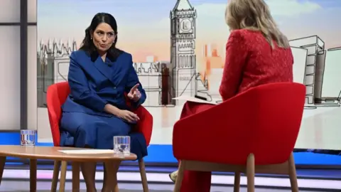 Dame Priti Patel, wearing a blue jacket and skirt, is sat in a red chair while talking to Laura Kuenssberg, who is also sat in a red chair and has her back to the camera.