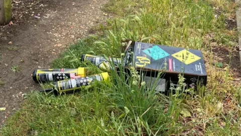 Discarded nitrous oxide canisters and packaging lying in long grass beside a footpath. Several black canisters with yellow labels are scattered next to a black box marked with hazard warning symbols.