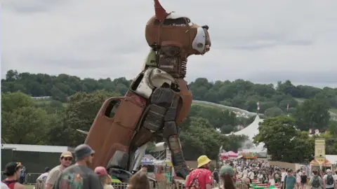 Crowds walk around a statue of a dog made from scrap cars and metal
