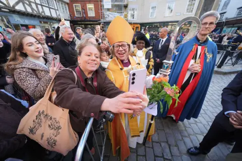 Yui Mok / PA Media The new archbishop takes a selfie with a woman at the front of a queue of people in Canterbury with village buildings in the background.