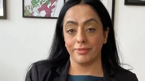 BBC Arooj Shah has long black hair and is wearing and black jacket with charcoal grey top.