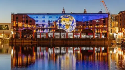 Adam Kenrick Huge projected images are reflected onto a brick building on the water. The images reflect back into the water in the evening's darkness. The projected pictures show a globe on a large blue backlight.