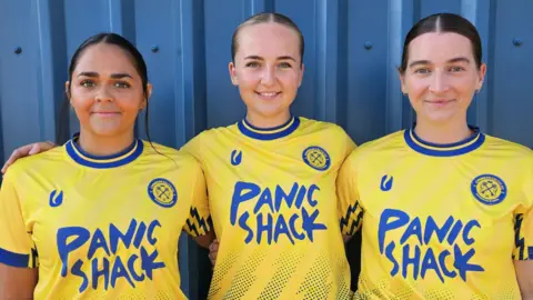 Matt Jarrett Three of the women's team players stand smiling at the camera in yellow home kit with blue sleeves and writing on the front