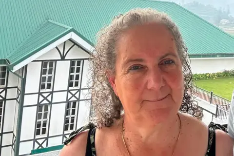 Alan Dutton A woman in her 60s with grey curly hair pulled back from her face stands in front and slightly above of a large white building with black window frames and a green roof. She is wearing and has tie straps on her shoulders. In the distance a misty hill side is in view.
