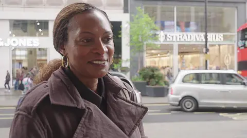 A woman in a brown coat stands on a high street in London. 