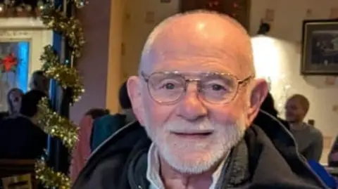 Gloucestershire Police A man in his 70s faintly smiles as he sits in a crowded restaurant, which has been decorated for Christmas, with tinsel wrapped around a pole. People can be seen sat around tables behind him.