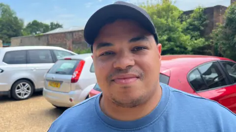 John Devine/BBC Dujon Pearman is standing in a shingled car park which has several cars parked on it. Pearman is wearing a black hat and a blue T-shirt.