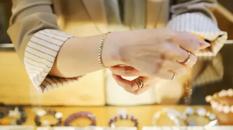 A woman with a striped brown and white shirt and pale blazer tries on a gold bracelet, with other bracelets on the table in front of her.