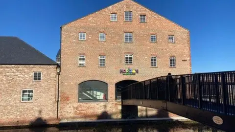 The brick building has been cleaned up. It has a series of small white windows above the first floor, and a black bridge over the river conects to the entrance.