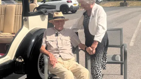 Peter Trotman (L) and Betty Trotman (R) by an old classic car. Peter is sitting in a chair and is wearing khaki trousers and a sun hat with a black band around it. Betty is wearing a black and white polka dot dress with a white jacket.