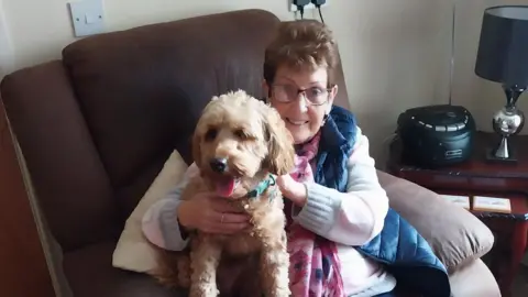 An older woman is sitting on a seat in a living room. She has short brown hair, wears glasses and a coat and is holding a dog.