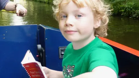 Kai wears a green T-shirt as they sit on a boat reading a Collins book about waterways 