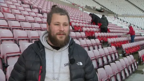 Chris Forsyth, co-founder of This Is Wearside, wearing a grey hoodie and black coat. Behind him are seats at stadium of light