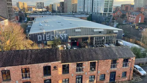 The Diecast complex as seen behind a set of redbrick terraced buildings, with the skyscrapers of Manchester's skyline seen in the distance.